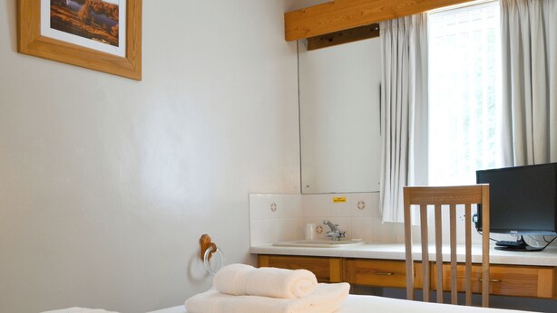 Una camera d'albergo con un letto, una sedia in legno, una scrivania con un computer, uno specchio e una finestra con tende.