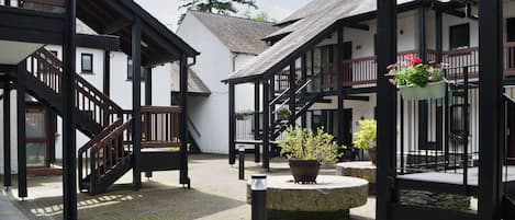 A courtyard with a stone fountain, wooden benches, and a staircase leading to a building with a balcony.