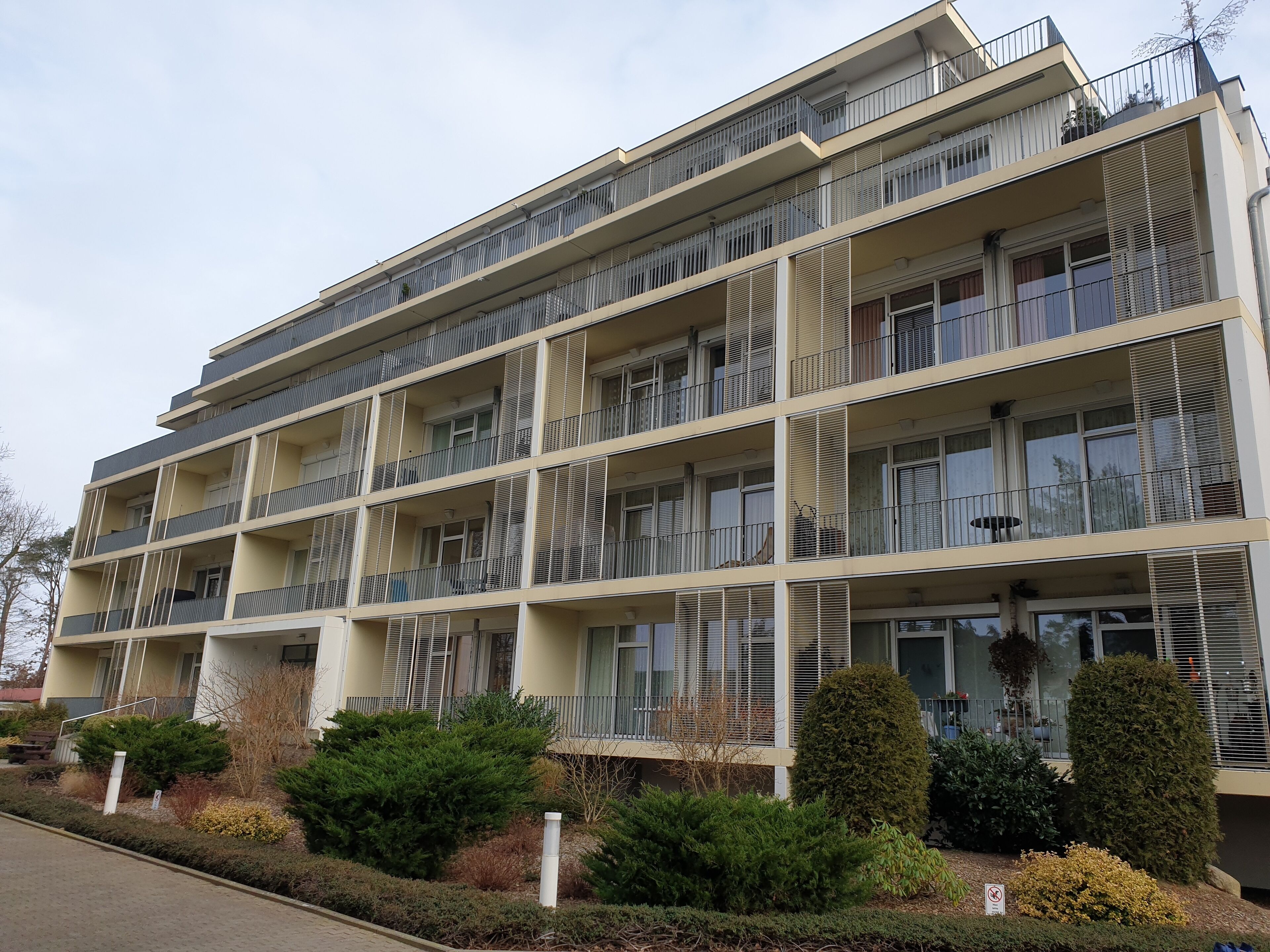 Ein mehrstöckiges Wohngebäude mit Balkonen, einer flachen Dachterrasse und einem Garten mit Sträuchern und einem kleinen Baum.