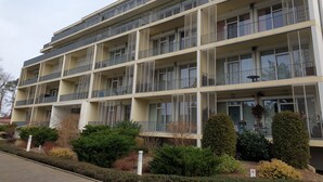 A multi-story residential building with balconies, a flat roof, and a garden area with shrubs and a small tree. - Excl. Apartment close to the beach, swimming pool, sauna, WLAN, free spa card, max. 3 pers. (Zempin)