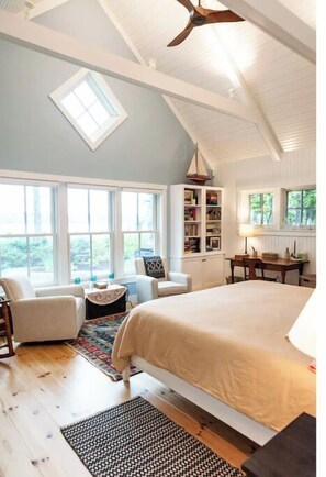 Relax in this serene bedroom with a large bed, ceiling fan, and a window offering a view of lush greenery.
