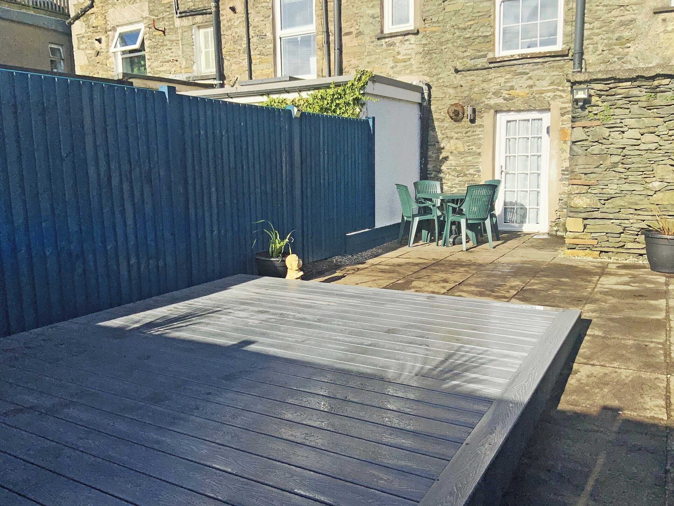 Une terrasse avec une table et des chaises en bois, une clôture bleue et un bâtiment en pierre en arrière-plan.