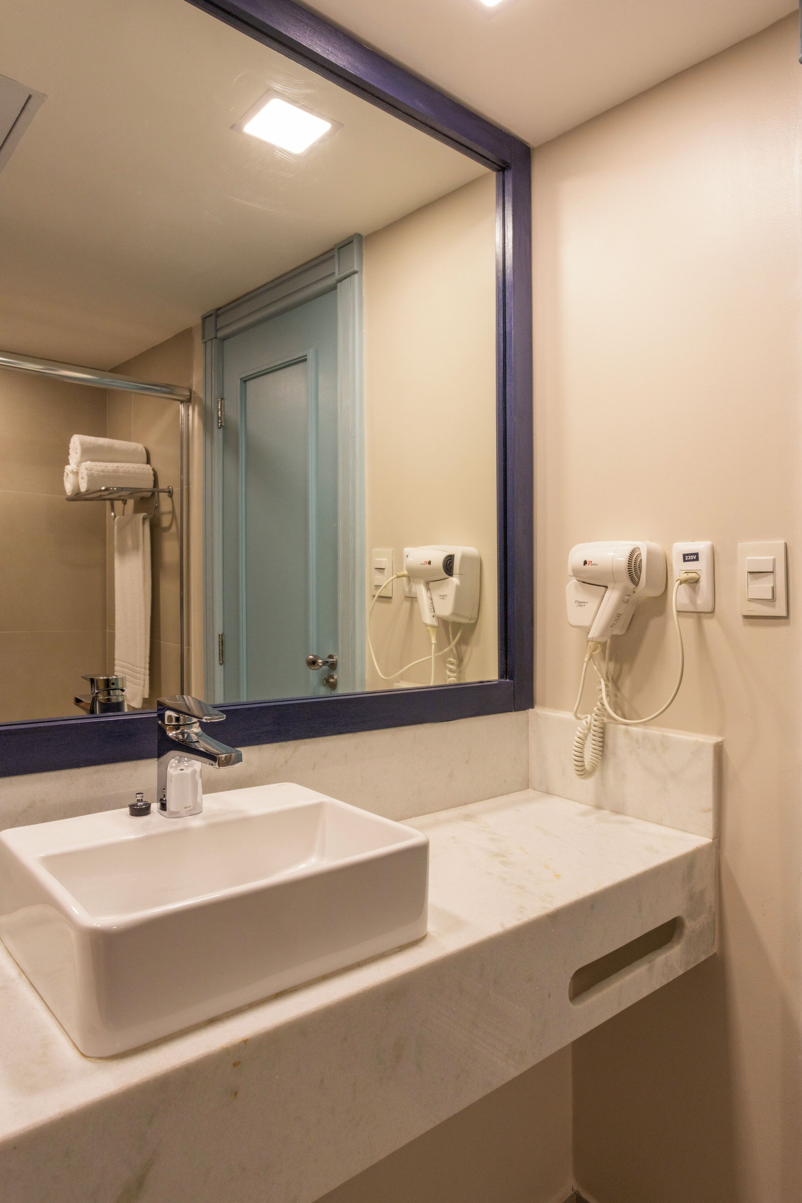 Une salle de bain avec un lavabo blanc, un miroir, une porte bleue et un sèche-cheveux fixé au mur.