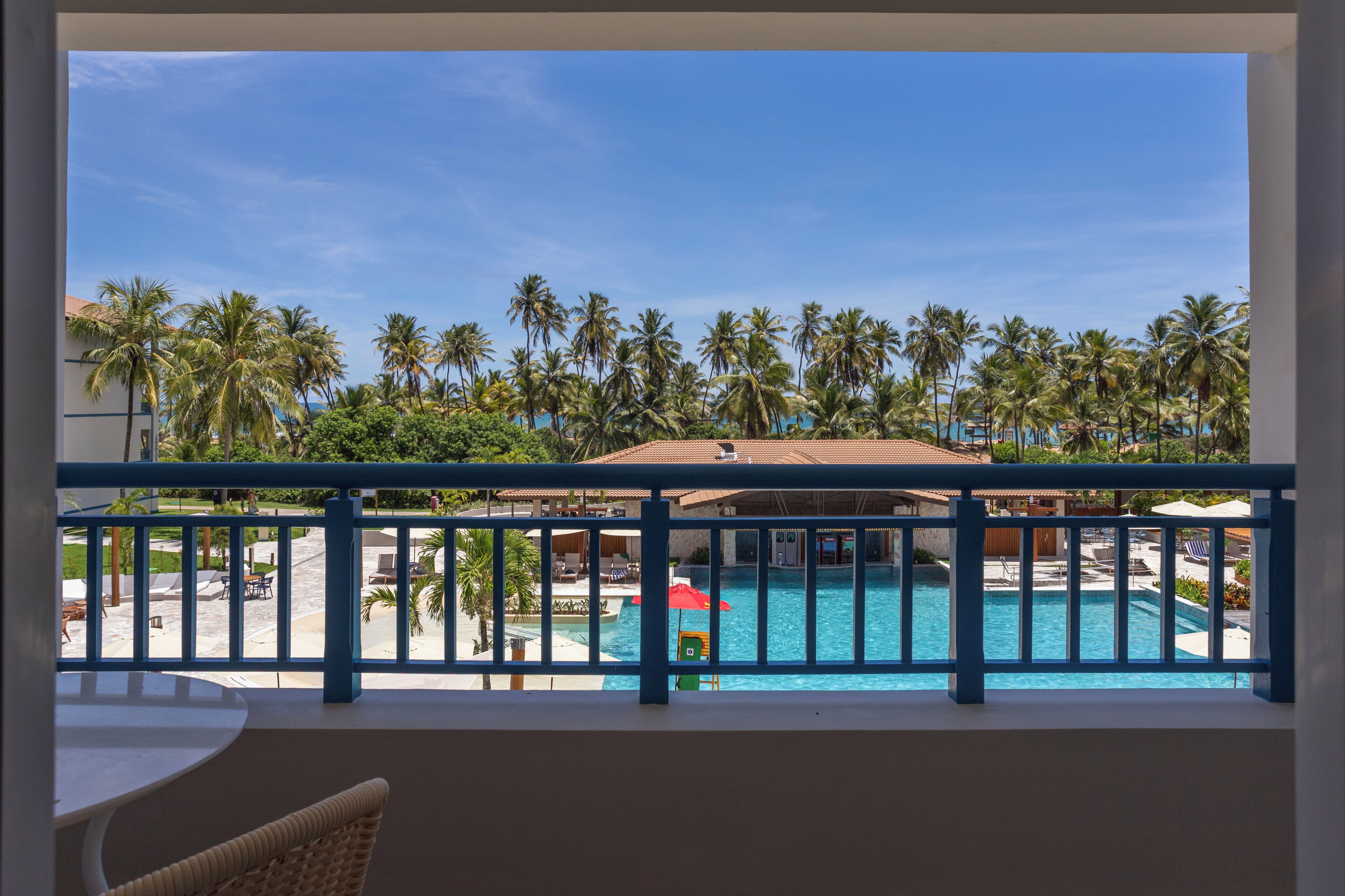 Uma varanda com vista para uma piscina, palmeiras e um edifício com telhado vermelho.