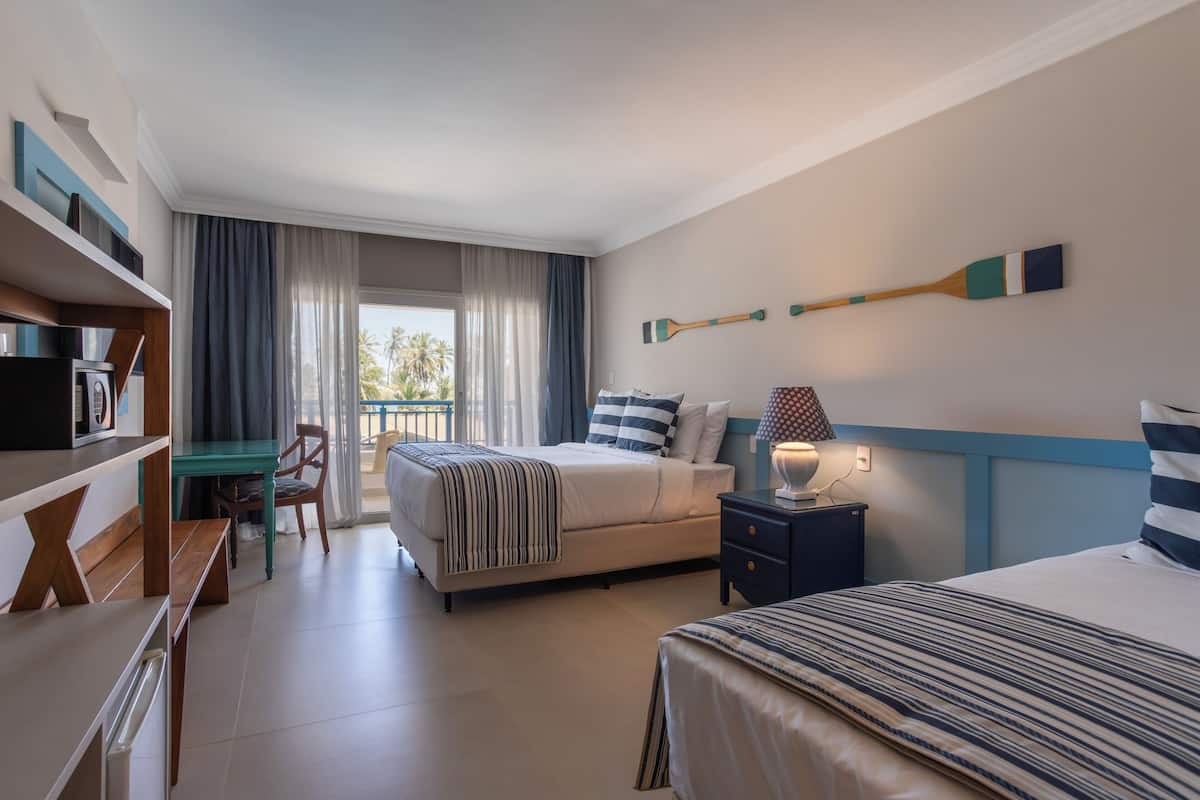 Une chambre d’hôtel avec un lit, un bureau, une chaise, une table de chevet et une vue sur des palmiers.