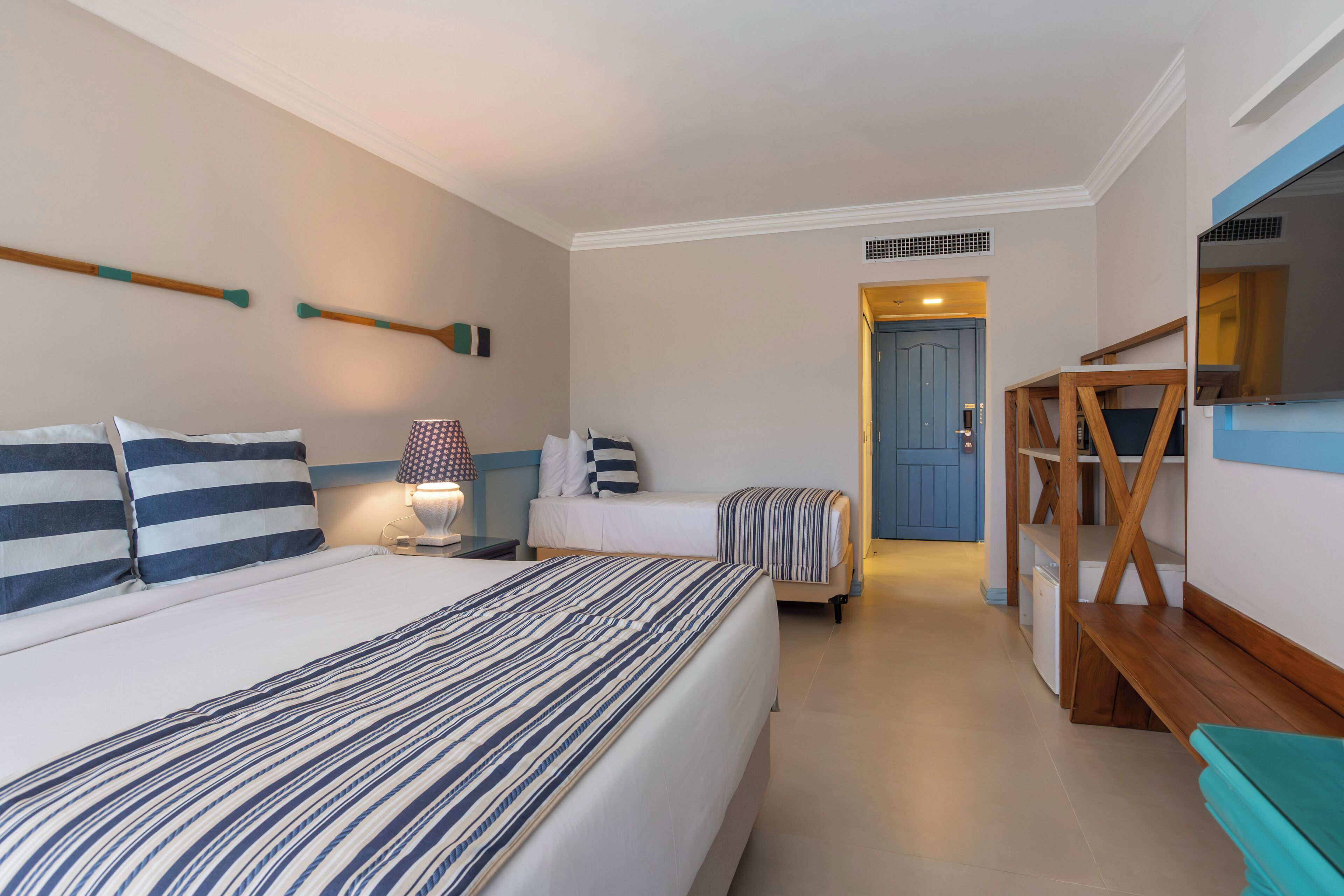 A hotel room with a large bed, a bedside table, a lamp, a small table, a bench, and a door leading to another room.