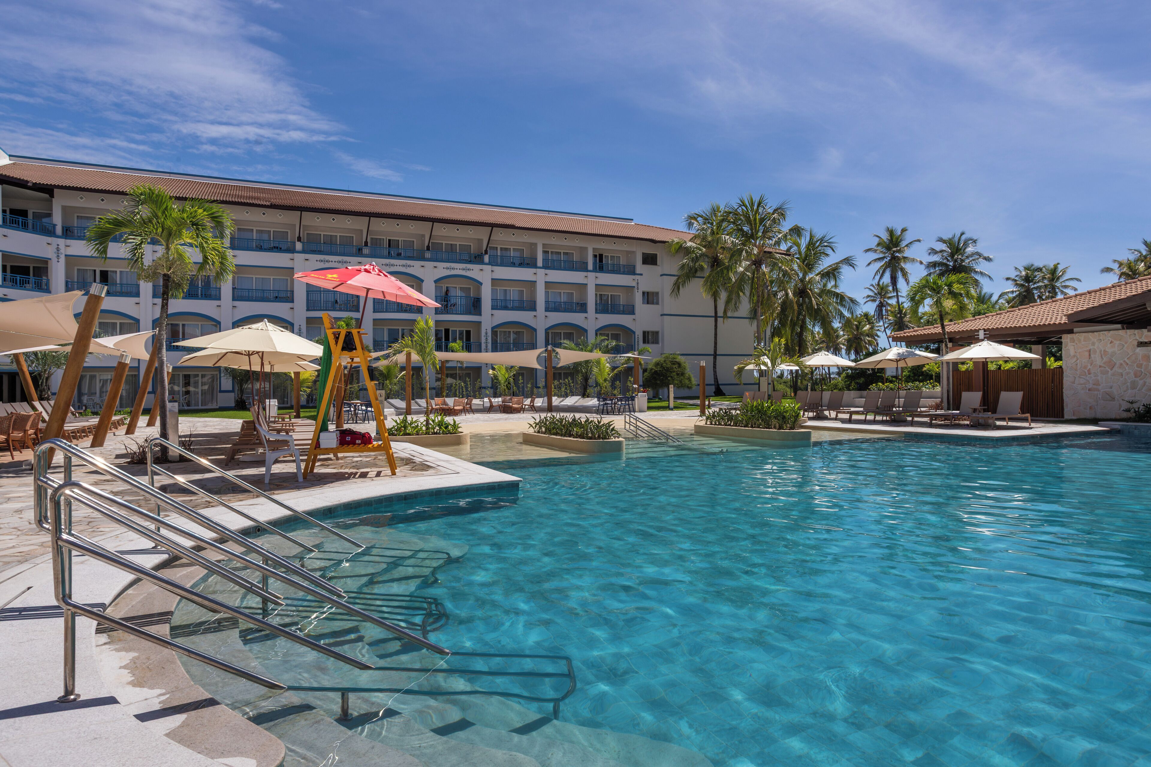 A hotel pool area with clear water, lounge chairs, umbrellas, and palm trees.