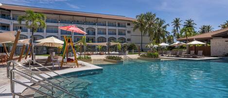 A hotel pool area with clear water, lounge chairs, umbrellas, and palm trees.