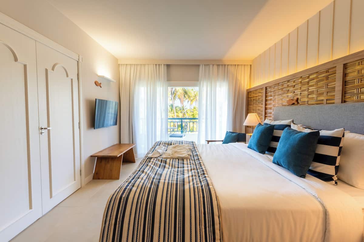 Une chambre d’hôtel équipée d’un lit, d’une télévision, d’un bureau et d’une fenêtre donnant sur des palmiers.
