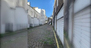 Une ruelle étroite pavée, bordée de bâtiments blancs à l’architecture traditionnelle, un ciel dégagé et un lampadaire bien visible.