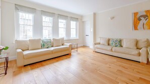 A spacious living room with two white sofas, a coffee table, and large windows with white curtains. - Spacious and Central 3 Bedroom House in Paddington (London)