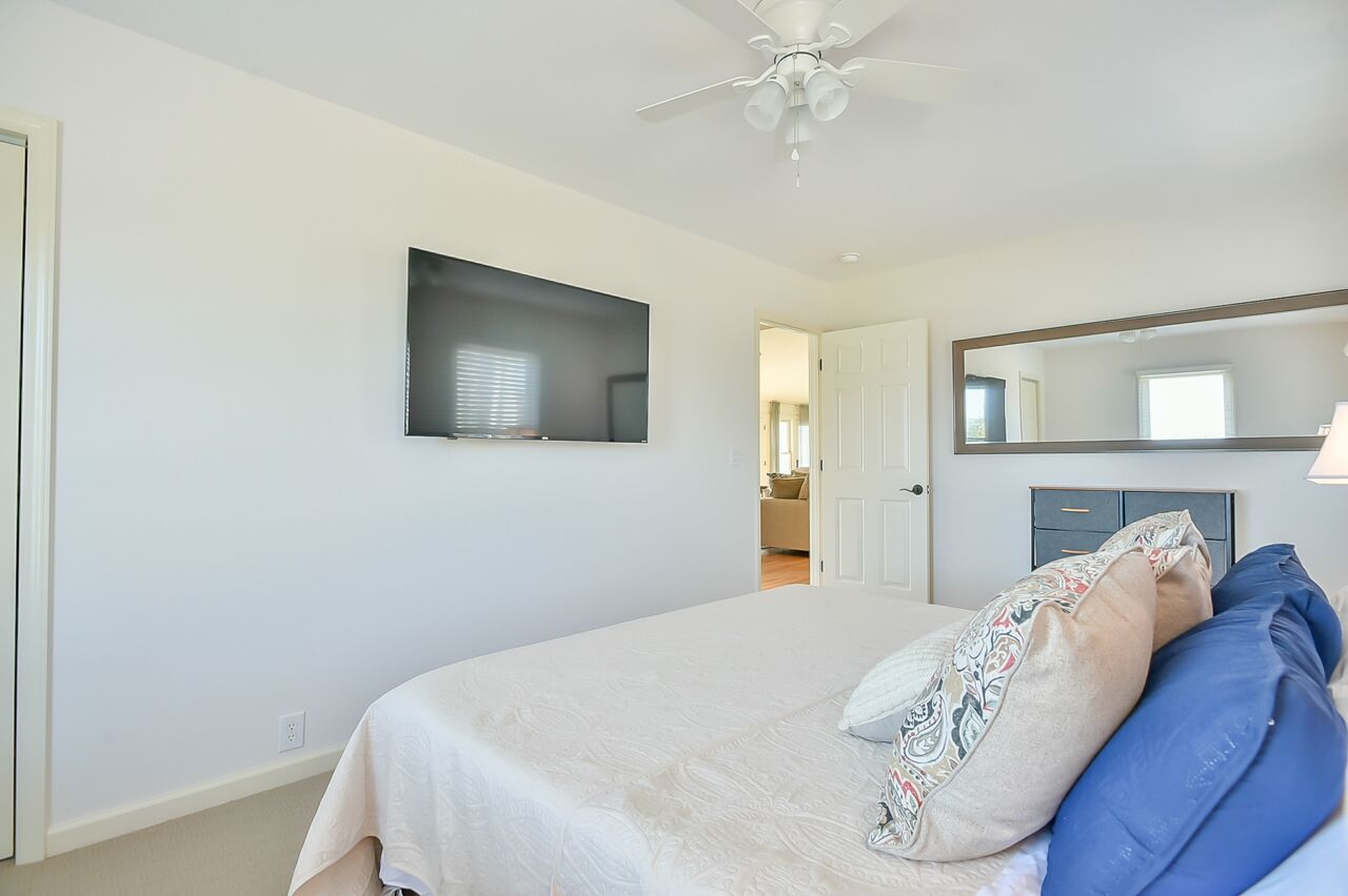 Une chambre à coucher comprenant un lit, un ventilateur de plafond, un téléviseur fixé au mur et une porte donnant sur une autre pièce.