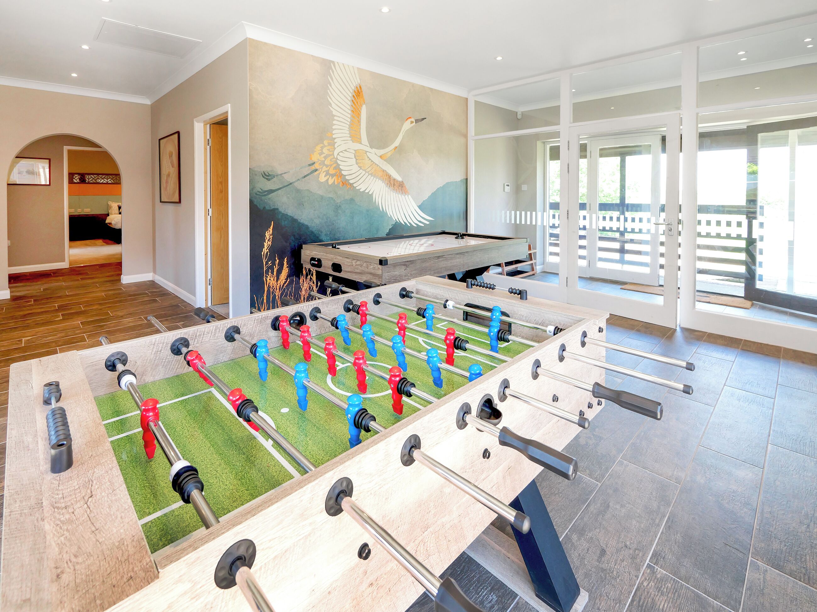 A foosball table in a room with a mural, large windows, and a wooden floor.
