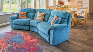 A modern living room with a blue sectional sofa, a dining area with wooden furniture, and large windows offering a view of greenery. - 4 bedroom accommodation in Carlton Colville, near Lowestoft (Carlton Colville, near Lowestoft)