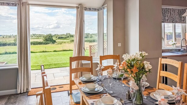 A dining area with a table set for four, chairs, a vase with flowers, and a view of a green landscape through large windows. - 4 bedroom accommodation in Carlton Colville, near Lowestoft (Carlton Colville, near Lowestoft)