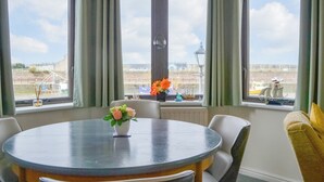 Une table ronde entourée de chaises, un vase rempli de fleurs et une vue sur la mer, visible à travers de grandes fenêtres ornées de rideaux.