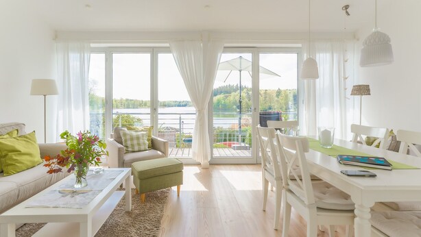 A bright living room with a sofa, a dining table, and chairs. There is a view of a lake through large windows. - Lakeview 2 Lake Salem child-friendly bathing lake W LAN (Salem)