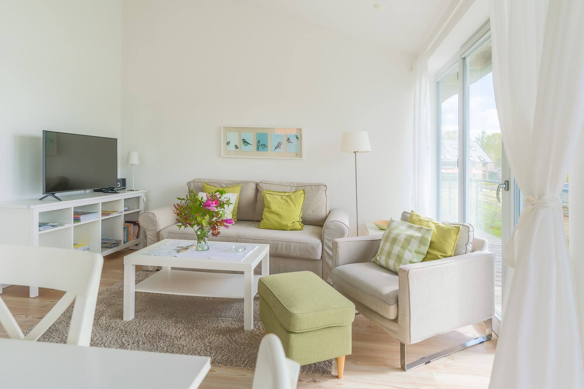 Een moderne woonkamer met een flatscreen televisie, een bijzettafeltje met een bloevas, een bank met kussens, en een boekenkast.