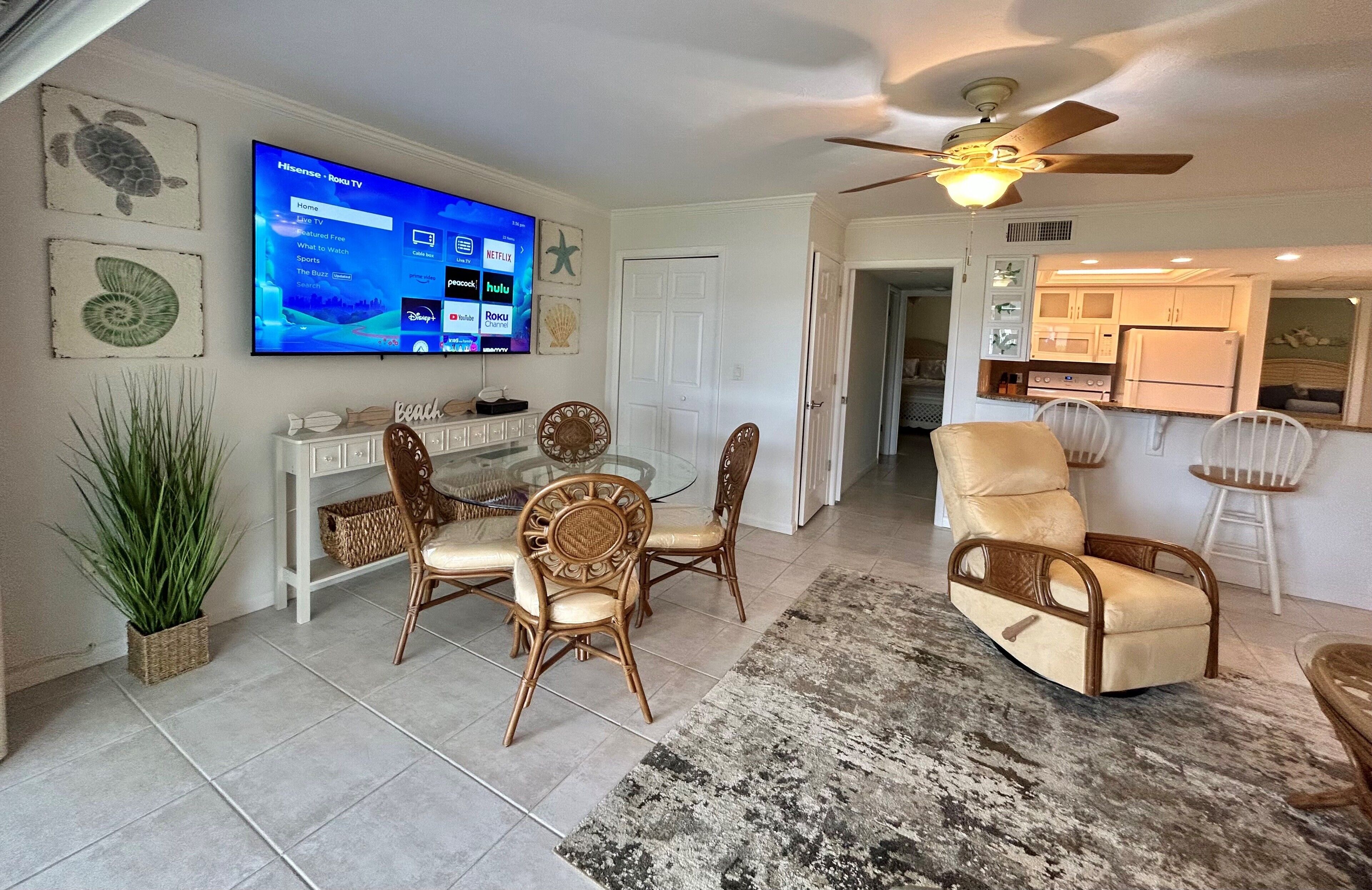 A living room with a TV, dining table, chairs, a ceiling fan, and a kitchen area.