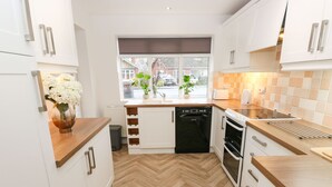 A modern kitchen with white cabinets, a herringbone wood floor, a dishwasher, an oven, and a window with plants. - Maple House (Tadcaster)