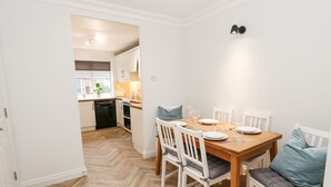 A dining area with a wooden table set for four, white chairs, and a kitchen in the background. - Maple House (Tadcaster)