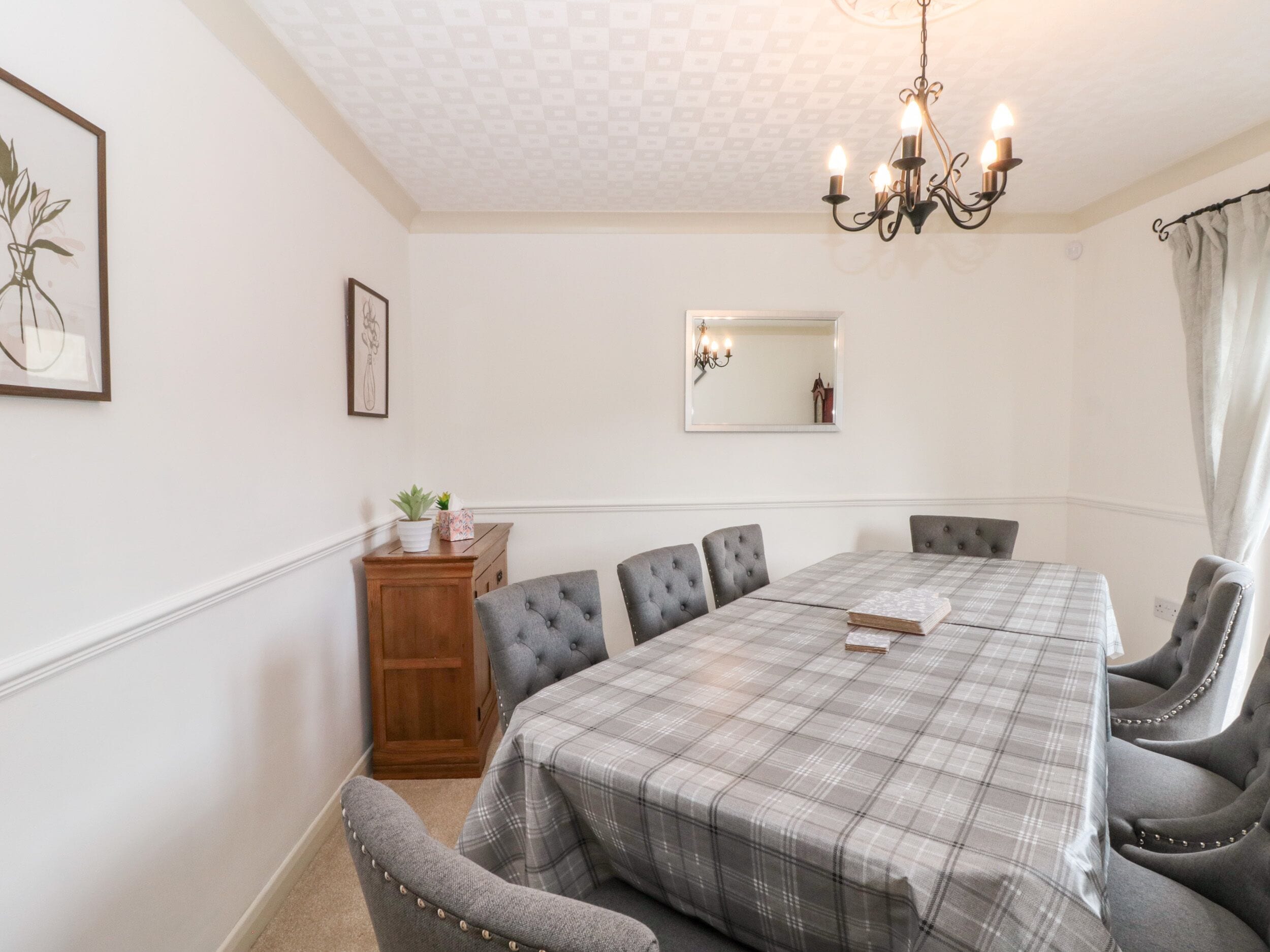 A dining room with a plaid tablecloth, grey upholstered chairs, a wooden sideboard, and a chandelier.