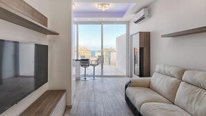 Una sala de estar moderna con televisor grande, sofá y balcón con vista a la ciudad.