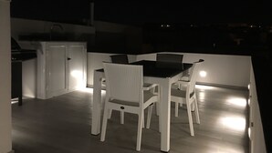 A patio area with a dining table and chairs, a small kitchen area with a refrigerator, and a view of the surrounding area at night.