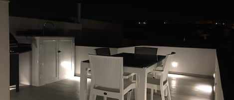 Un patio con mesa y sillas, un pequeño espacio de cocina con refrigerador, y vistas del entorno nocturno.