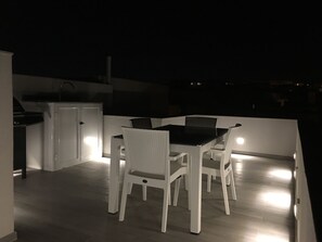 Un patio con mesa y sillas, un pequeño espacio de cocina con refrigerador, y vistas del entorno nocturno.