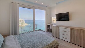 Un dormitorio con una cama grande, un televisor montado en la pared, un escritorio con una silla y vistas al océano y edificios a través de la ventana.