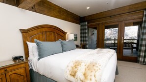 A bedroom with a wooden headboard, a large bed with a white and blue bedding set, a bedside table with a lamp, and a view of a snowy landscape through a glass door. - Deluxe King Room With Deck Hotel Room by RedAwning (Park City)