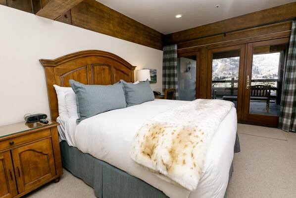 A bedroom with a wooden headboard, a large bed with a white and blue bedding set, a bedside table with a lamp, and a view of a snowy landscape through a glass door. - Deluxe King Room With Deck Hotel Room by RedAwning (Park City)