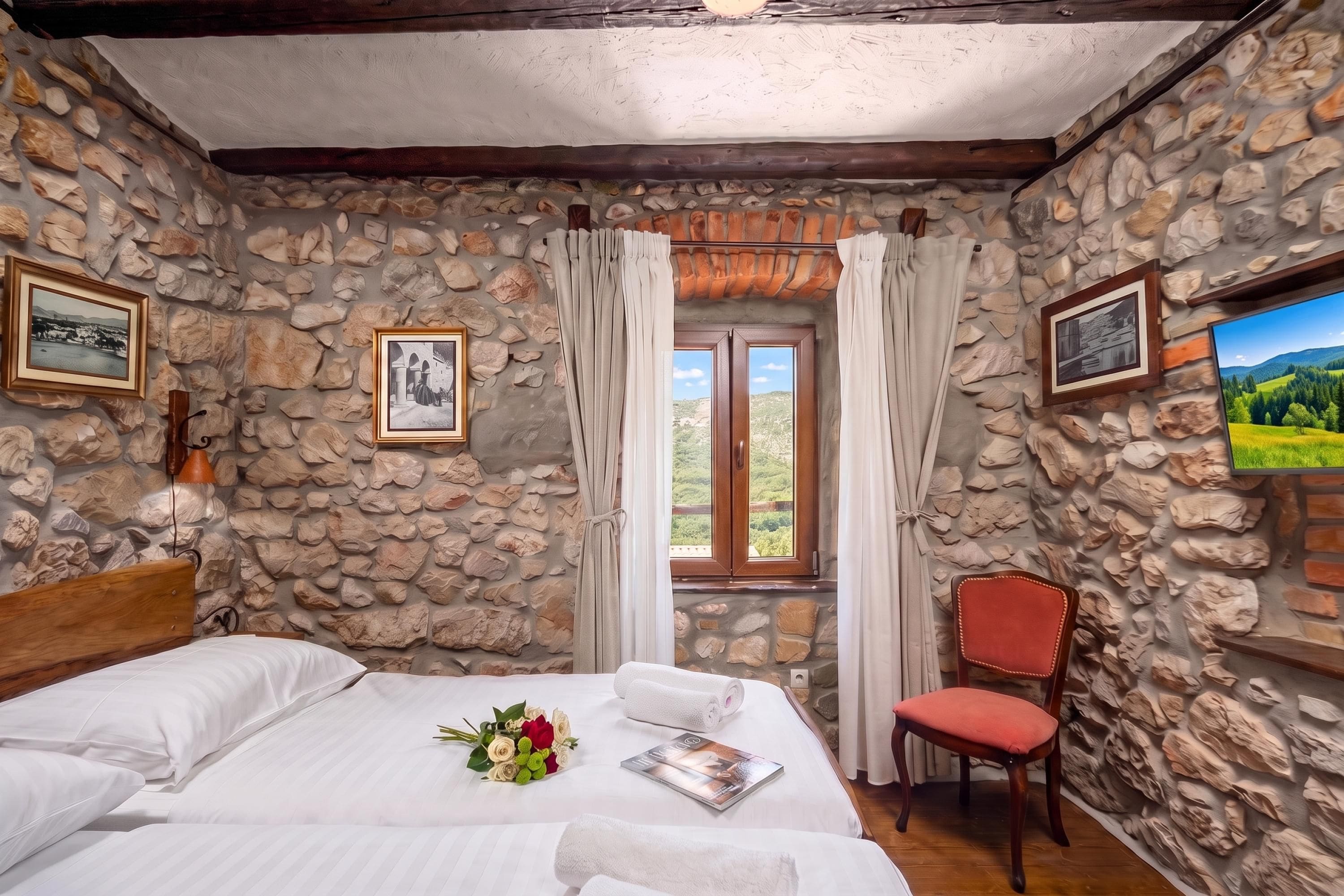 Une chambre avec des murs en pierre, un lit en bois, une chaise rouge et une fenêtre avec des rideaux.