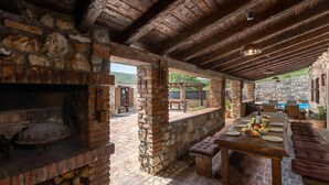 An outdoor patio with a stone fireplace, wooden beams, a dining table with plates and fruits, and a pool in the background. - Villa Cupido With Private Pool (Vinodol)