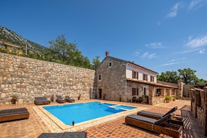 Un espace piscine entouré de murs en pierre, avec des chaises longues et un bâtiment en pierre doté d’un toit en tuiles.
