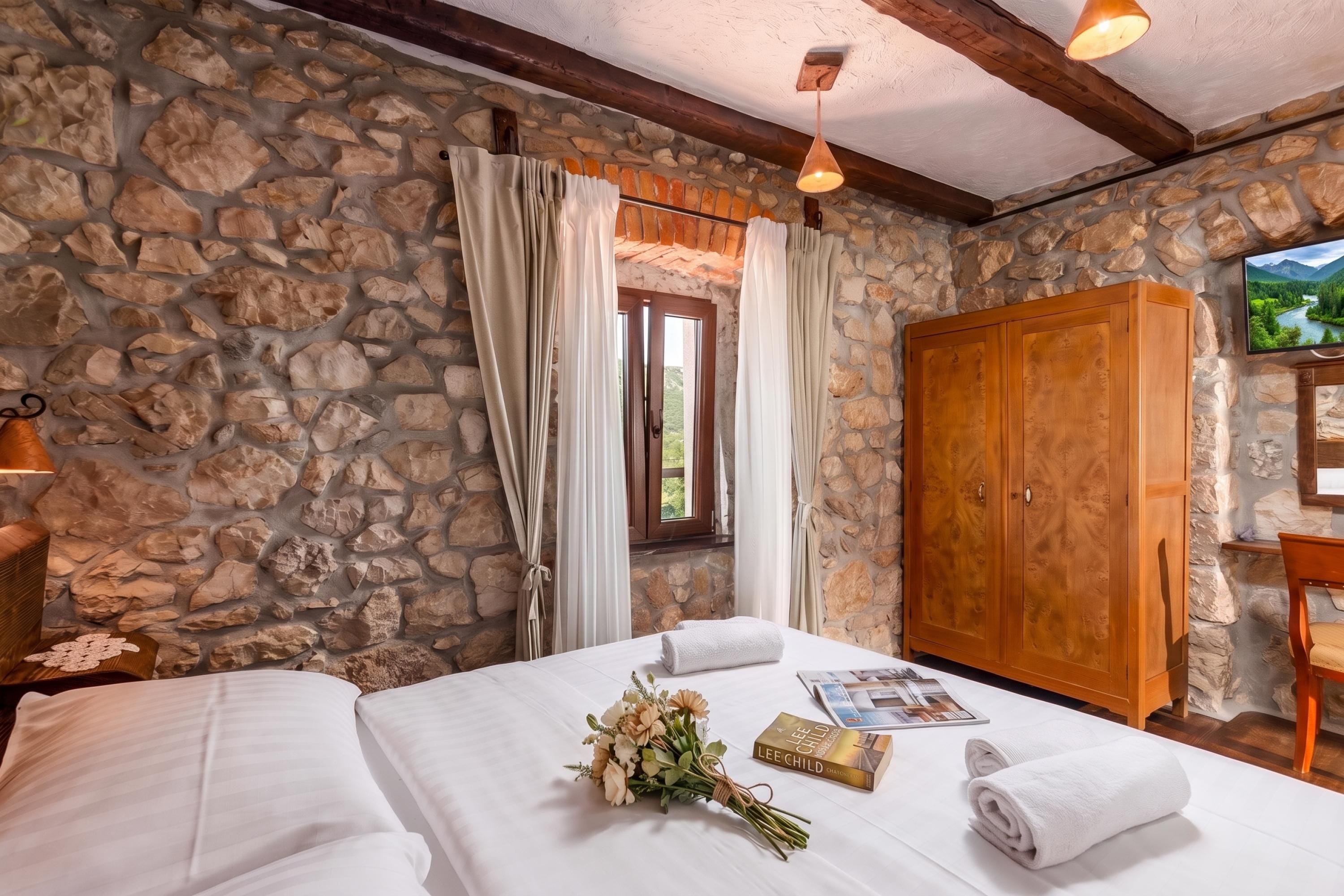 Un dormitorio con paredes de piedra, un armario de madera, una cama con ropa blanca, una ventana con cortinas y un televisor en la pared.