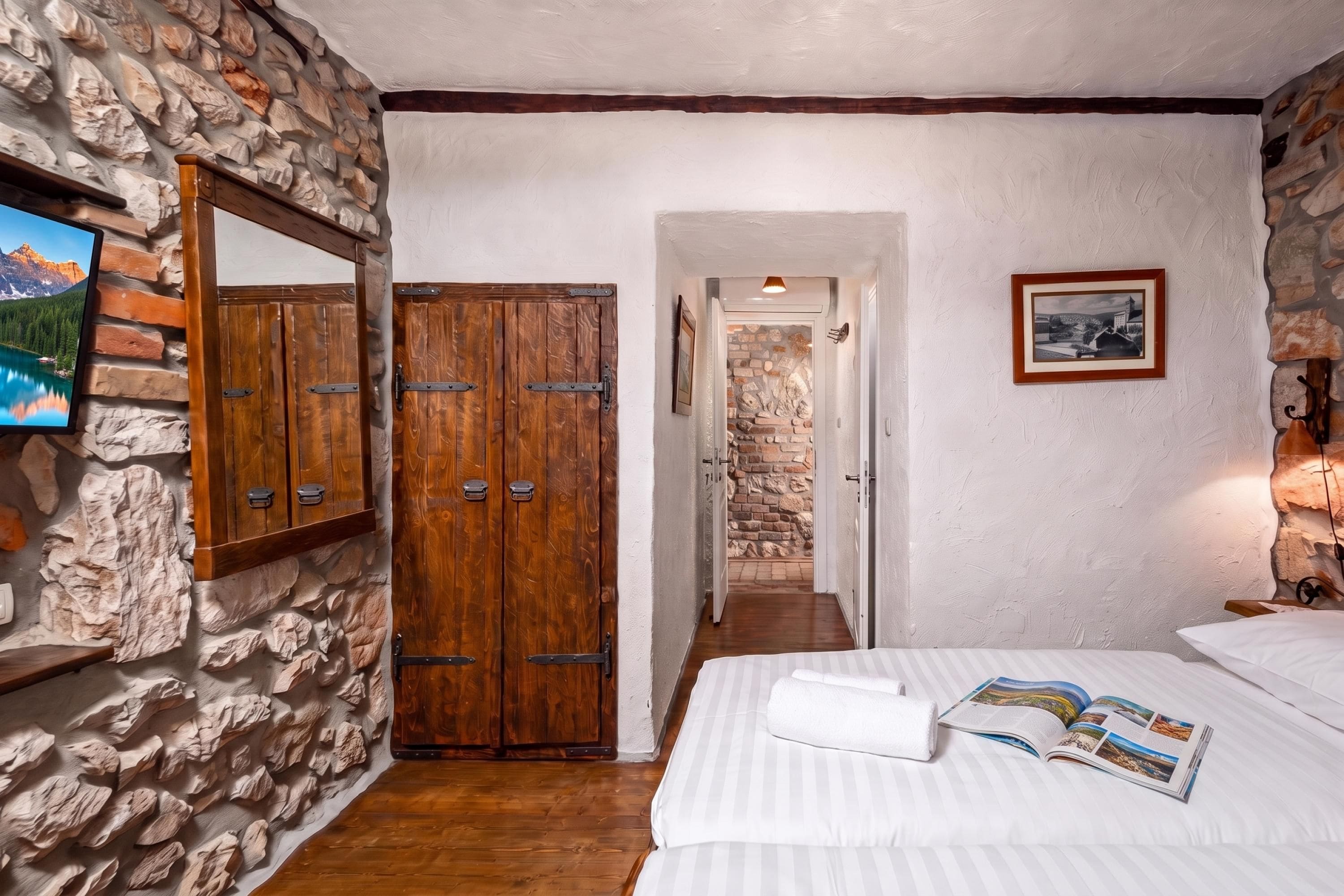 A room with a stone wall, wooden wardrobe, bed with white bedding, and a framed picture on the wall.