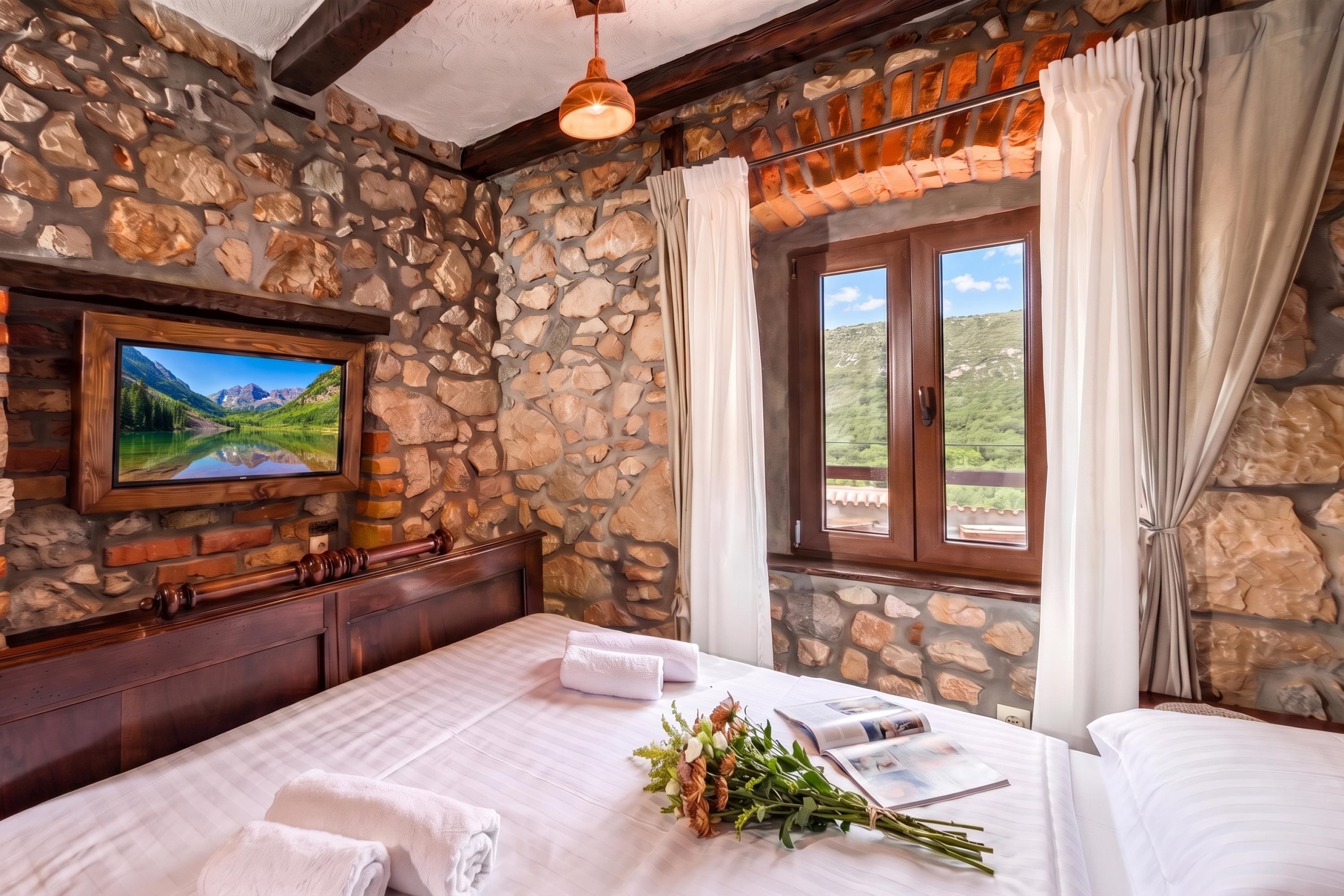 A room with stone walls, a bed with white linens, a window with curtains, and a painting of a landscape.