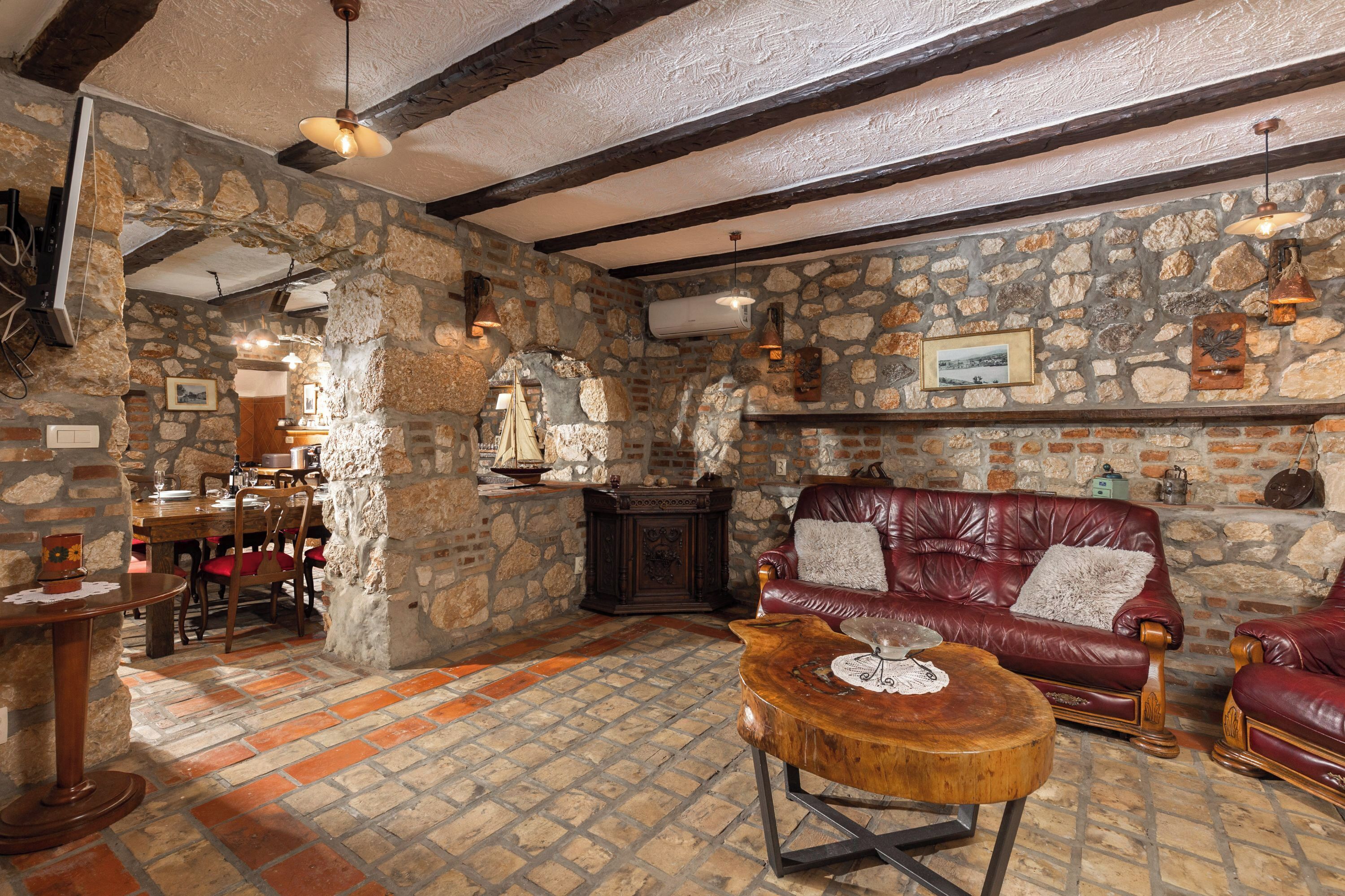 A stone-walled room with a wooden ceiling, a leather sofa, a wooden coffee table, and a dining area with chairs and a table.