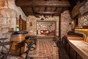 A rustic interior with stone walls, wooden beams, and brick flooring. There are tables and chairs, a fireplace, and a wooden barrel.