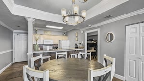 A dining area with a round table and white chairs, a kitchen in the background, and a chandelier. - Ocean Ritz-501 4 Bedroom Condo by RedAwning (Panama City Beach)