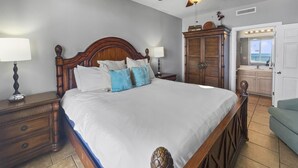 A neatly arranged bedroom with a large bed, wooden headboard, two bedside tables with lamps, a wardrobe, and a bathroom visible through an open door. - Ocean Ritz-501 4 Bedroom Condo by RedAwning (Panama City Beach)