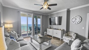 A living room with a ceiling fan, a flat-screen TV, a clock, and a view of the ocean through large sliding doors. - Ocean Ritz-501 4 Bedroom Condo by RedAwning (Panama City Beach)