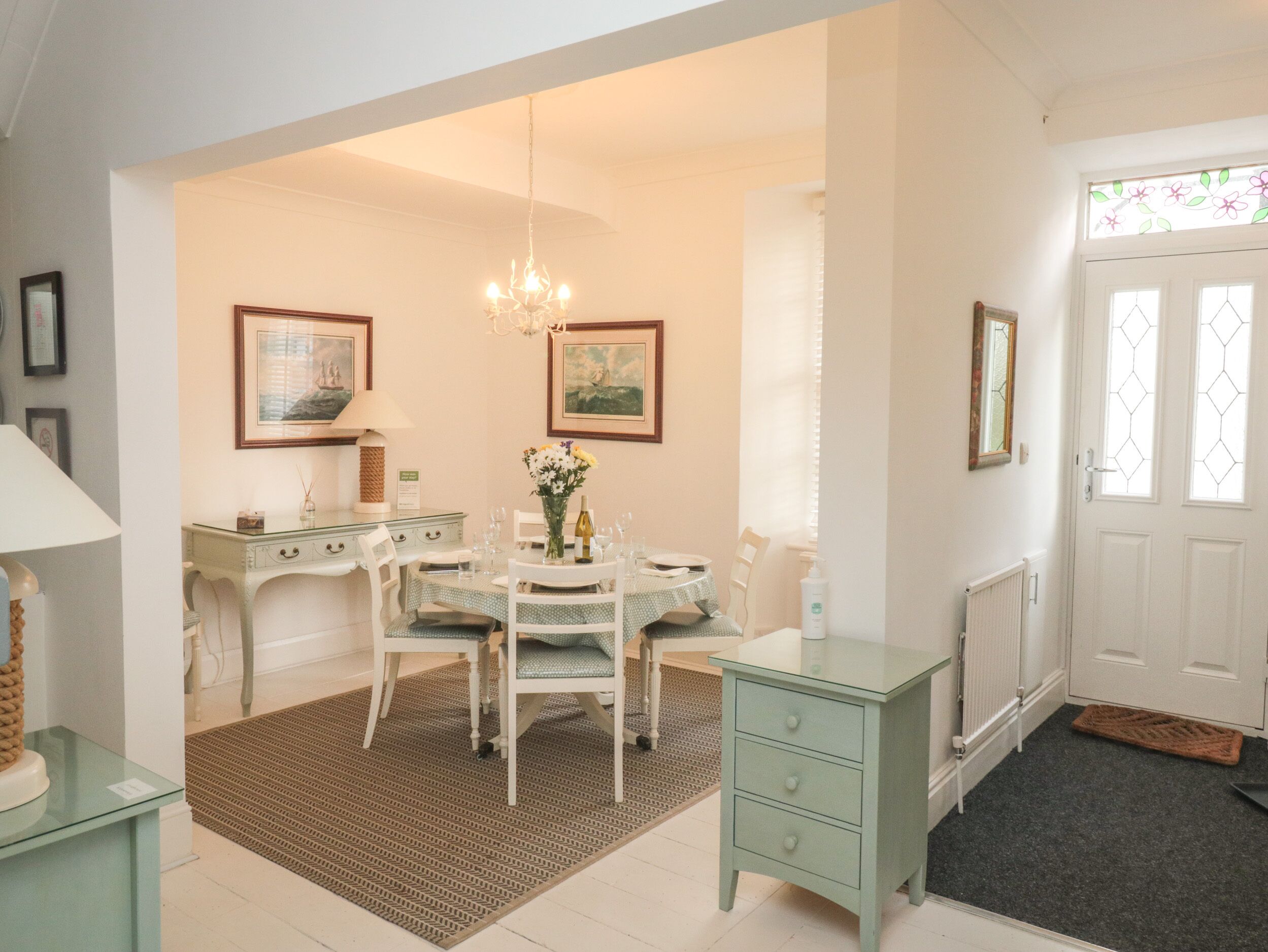 A dining area with a table set for four, a chandelier, and two framed paintings on the wall.