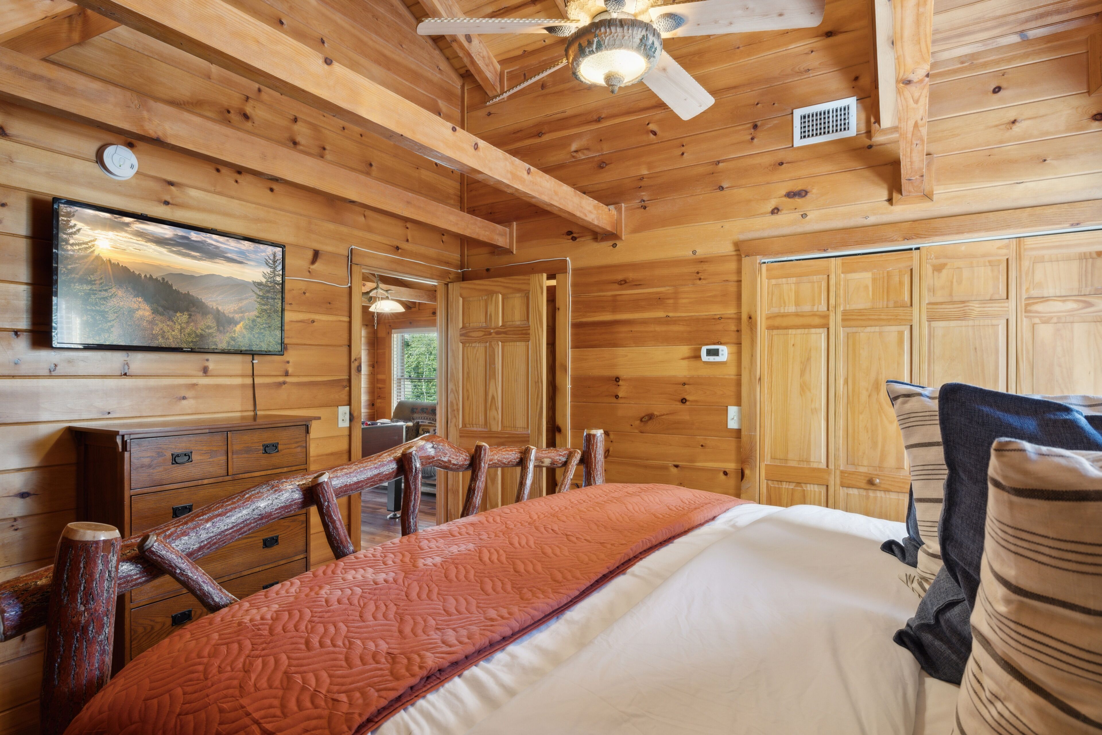 Una habitación de cabaña de madera con una cama, un televisor montado en la pared y un ventilador de techo.