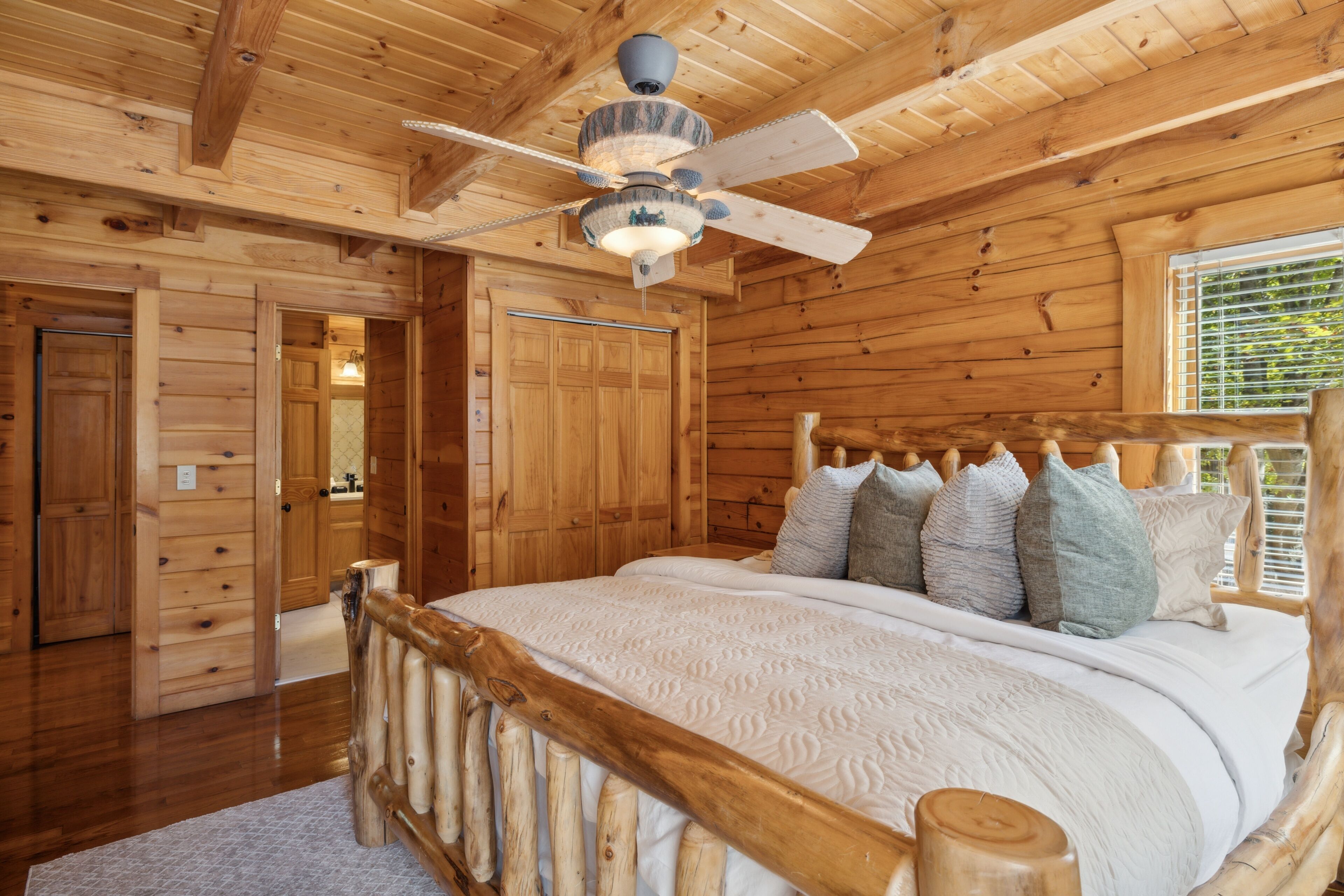 Dormitorio de cabaña de madera con cama, ventilador de techo de madera y ventana con persianas.