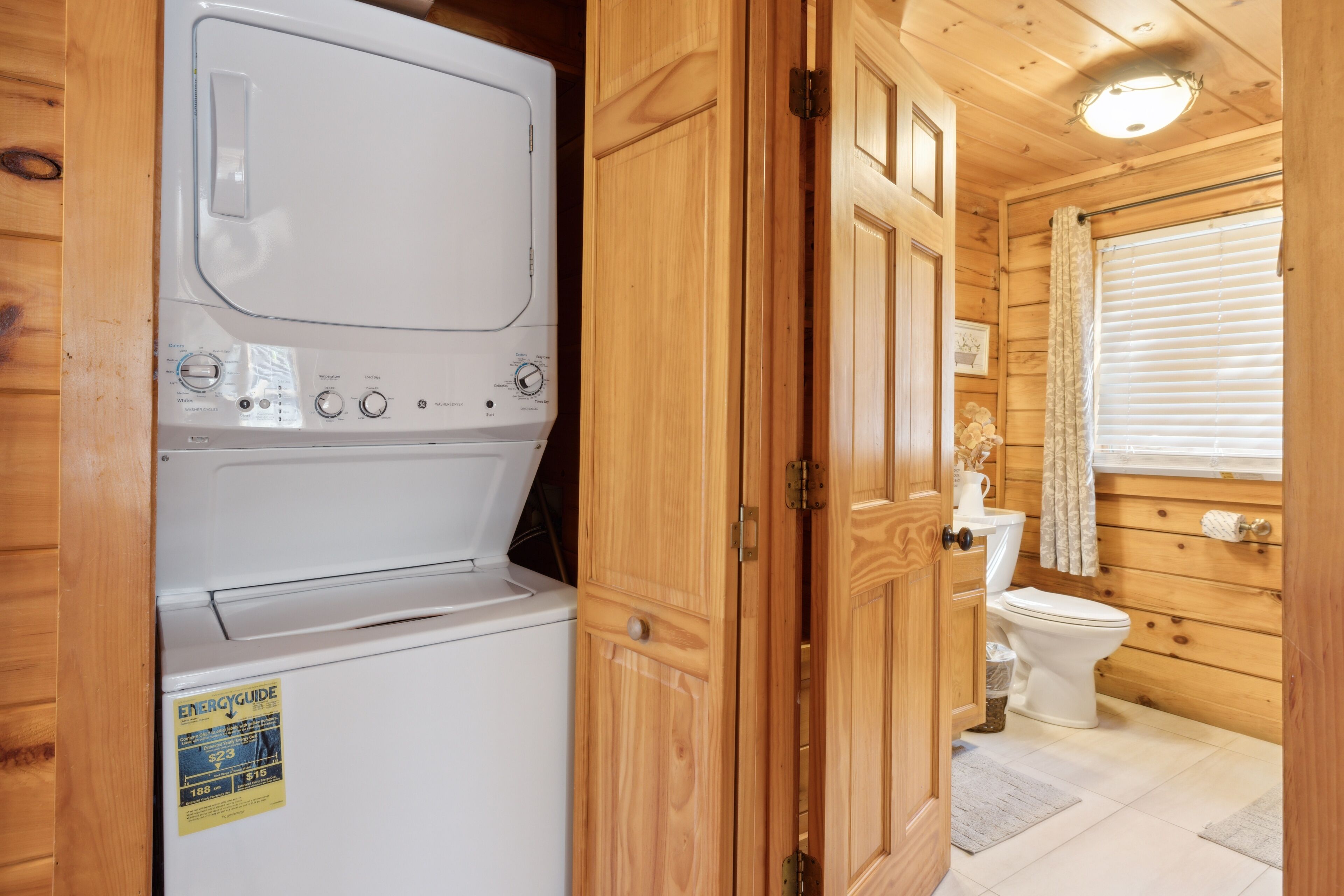 A compact, wooden cabin-style bathroom with a white washing machine, a toilet, and a window with blinds.