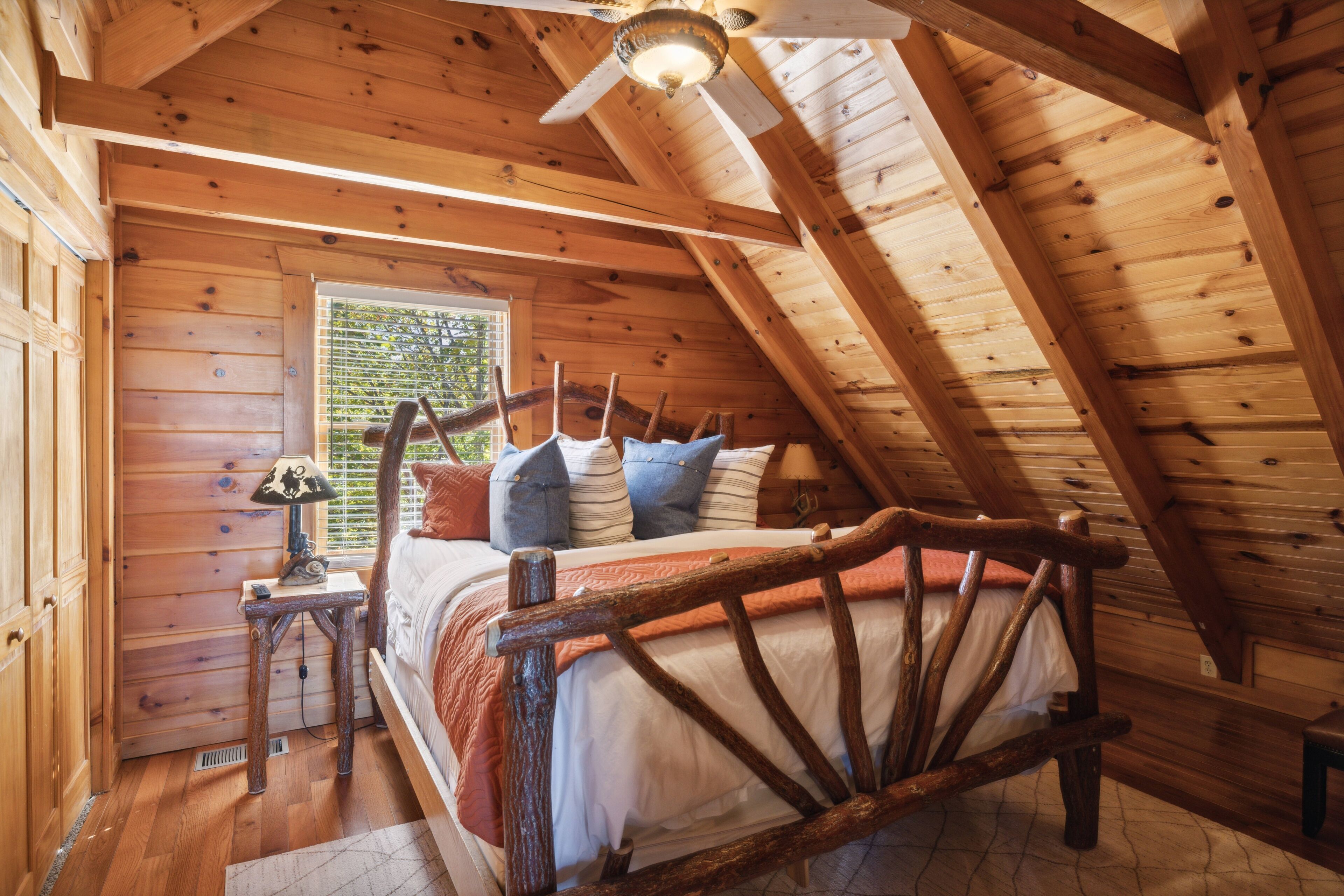 A wooden cabin bedroom with a sloped ceiling, a bed with a wooden headboard, and a window with blinds.