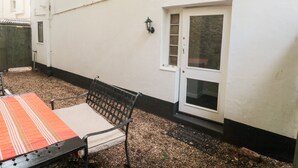 Een wit gebouw met een deur en ramen met zwarte kozijnen, een bank met een gestreept kussen en een klein tuinperk met grind.
